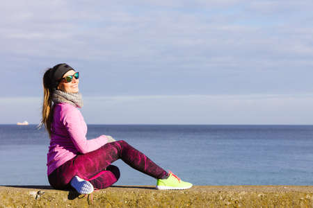 Woman resting relaxing after doing sports outdoors. Fitness girl female jogger wearing warm sporty clothes in cold day weather on seaside taking breakの写真素材