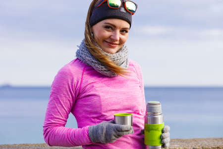 Woman athlete fit girl wearing warm sporty clothes outdoor in cold weather drinking hot tea from vacuum flask thermos, warming up. Sports, activities, hiking in winter or autumn timeの写真素材