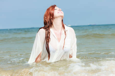 Vacation. Sensual girl wet cloth in water on the coast. Redhair woman having fun relaxing on the sea. Summertime.の写真素材