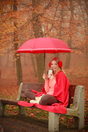 Nature outdoor weather fun cozy concept. Girl sitting in park. Lady sovering herself from rain by holding umbrella drinking coffee from cup while staying on bench.の写真素材