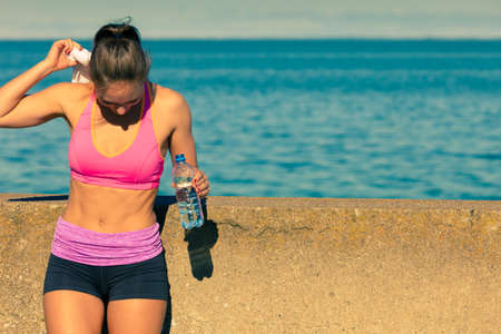 Woman in sportswear takes a break to rehydrate drinking water from plastic bottle, resting after sport workout outdoor by seasideの写真素材