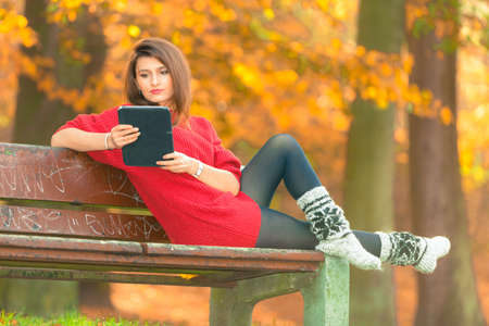 Technology and education. Woman relaxing in autumnal park sitting on bench with tablet. Pretty fashionable female student learn on fresh air.の写真素材