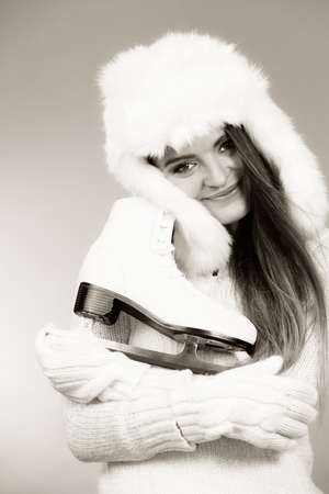Winter sport activity concept. Girl wearing warm hat and furry waistcoat holding and hugging ice skate, black and white studio shot.の写真素材