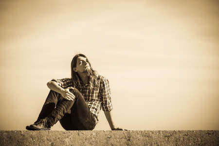 Man long hair wearing plaid shirt relaxing outdoor sitting on concrete wall at sunny day against skyの写真素材