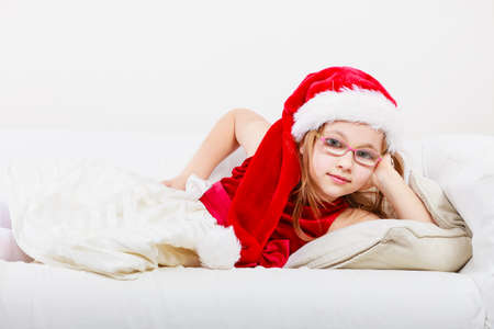 Christmas holiday concept. Toddler girl wearing Santa Claus hat and christmassy dress lying on sofa with pillows, cheek propped up by hand.の写真素材
