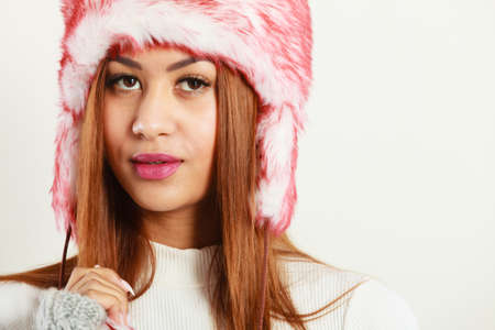 Winter clothing, fashion concept. Closeup of young mulatto woman wearing red white fur cap. Mixed...