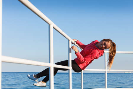 Outdoor, dangerous play, sport concept. Woman in sports suit sitting on handrail next to sea and having fun while exercising.の写真素材