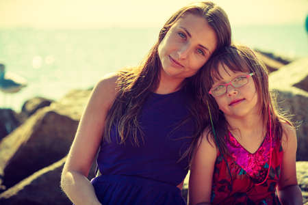 Mother and daughter posing near sea rock with water in background on sunny day