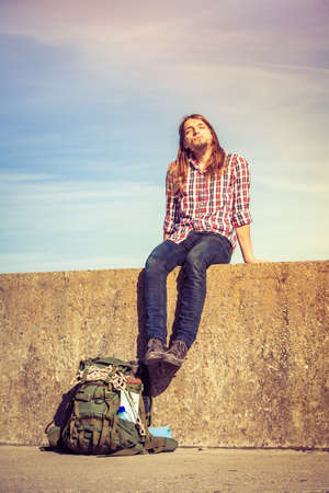 Man tourist backpacker relaxing outdoor sitting on grunge wall against sky. Adventure, summer, tourism active lifestyle. Young hipster guy trampingの写真素材