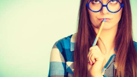 Studying, education and fun concept. Nerdy thinking woman in weird big glasses holding pen. Studio shot on blue backgroundの写真素材