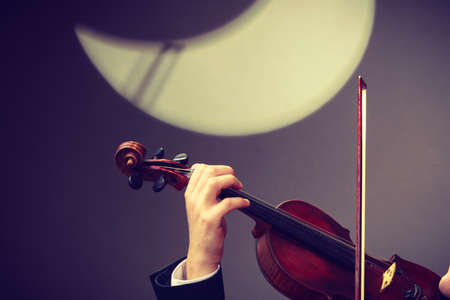 Music passion, hobby concept. Young man man dressed elegantly playing on wooden violin. Studio shot on dark background with white moonの写真素材
