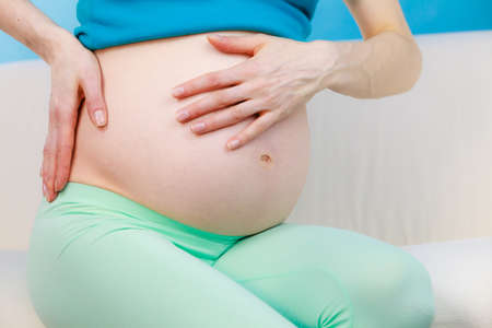Pregnant woman sitting on sofa at home with hands on her back, making massage, suffering from pain....