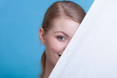 Workout, hygiene concept. Smiling woman holding a big white clean towel and hiding behind it, studio shot on blue backgroundの写真素材