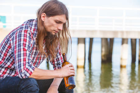 Man depressed with wine bottle sitting on sea shore outdoor. People abuse and alcoholism problemsの写真素材