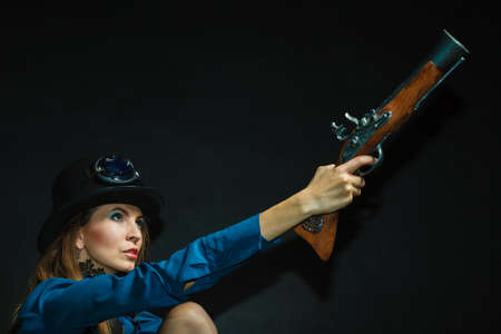 Subculture fashionable victorian elegant weapon concept. Steampunk girl armed and dangerous. Lady dressed in victorian fashion holding antique firearm aiming.の写真素材
