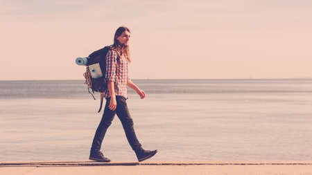 Man hiker backpacker walking with backpack by seaside at sunny day. Adventure, summer, tourism active lifestyle. Young long haired guy trampingの写真素材