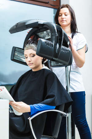 Haircare, relaxation and hairstyling concept. Woman sitting in black cape getting her hair dried under machineの写真素材