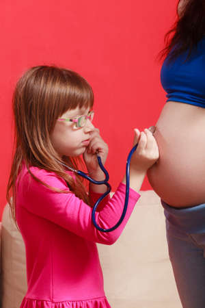 Pregnancy, family concept. Daughter using stethoscope on her pregnant mother belly to hear sibling heart tonesの写真素材