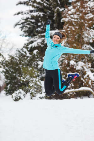 Sporty girl jumping in snow with trees in background. Winter sports, outdoor fitness, nature workout, health concept.の写真素材