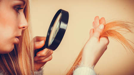 Haircare, health problem concept. Unhappy blonde woman looking at ends of her blonde hair through magnifying glassの写真素材