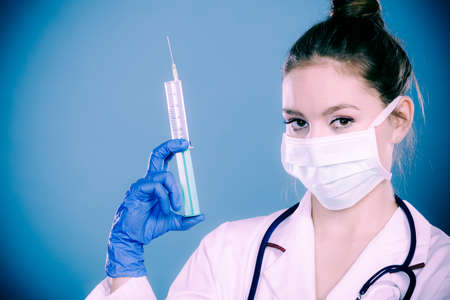 medication and cure injection concept young woman doctor with stethoscope holding syringe female specialist with mask on face and vaccine in handの写真素材