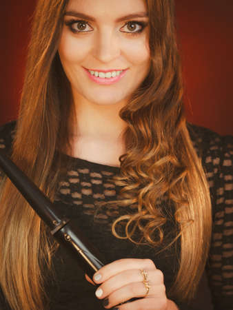 Haircare, hairstyiling at home concept. Portrait of young happy woman curling her long brown straight hair with curler. Studio shot on dark colorful backgroundの写真素材