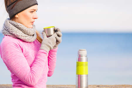 woman athlete fit girl wearing warm sporty clothes outdoor in cold weather drinking hot tea from vacuum flask warming up sports activities hiking in winter or autumn timeの写真素材