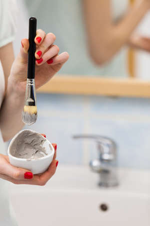 Skin care. Female hand holding brush and clay mud mask to apply on skin face, in bathroom. Girl taking care of her oily compexion. Spa wellbeing.の写真素材