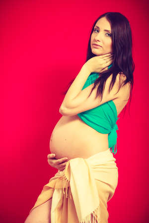 Parenthood, waiting for baby concept. Adult woman showing her big pregnant belly. Indoor shot on red backgroundの写真素材