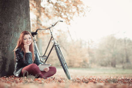 Outdoor nature fitness internet technology concept.Girl under tree with bike. Young redhead lady resting after cycling browsing websites on tablet.の写真素材