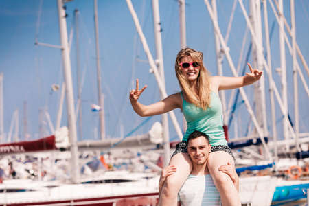 Relationship goals, summer love concept. Man giving girlfriend piggyback ride on marina, woman showing peace gesture.の写真素材