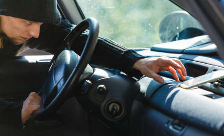 Anti theft system problem concept. Burglar thief man wearing black clothes breaking into car and stealing smartphoneの写真素材