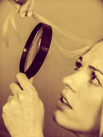 haircare health problem concept unhappy woman looking at ends of her blonde hair through magnifying glass sepiaの写真素材