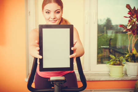 Active young woman working out on exercise bike stationary bicycle holding pc tablet computer with...