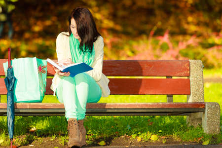 Woman enjoying beautiful autumn nature weather, reading book sitting on bench in park during sunny autumnal day,の写真素材