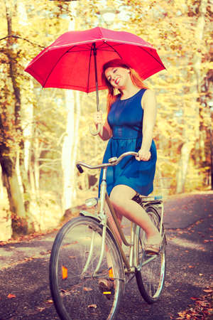 Nature, people, outdoors concept. Ginger hair girl is holding red umbrella. Lady is riding a bike and wearing blue dress.の写真素材