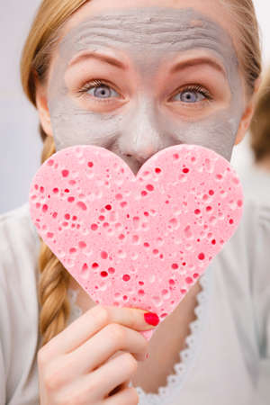 Facial dry skin and body care, complexion treatment at home concept. Happy young woman loving having grey mud mask on her face, holding sponge in heart shape.の写真素材