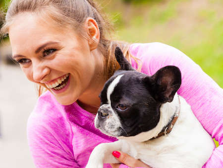 Animal lover, little pets, dogs fun concept. Happy smiling woman wearing sport clothes holding small cute French Bulldog in park outsideの写真素材