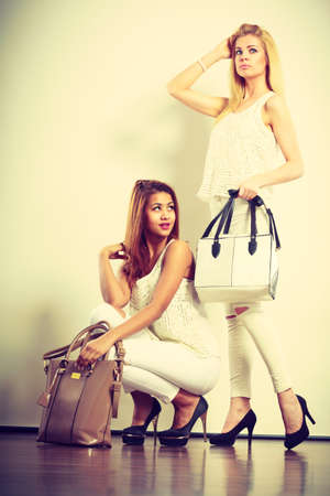 Fashion concept. Two women mulatto and blonde caucasian female in white fashionable clothes high heels shoes with bags handbags.の写真素材