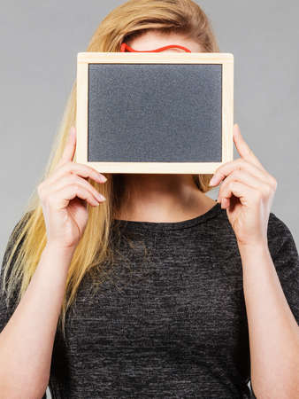Woman holding blank black board on face, hiding behind copyspace, grey background.の写真素材