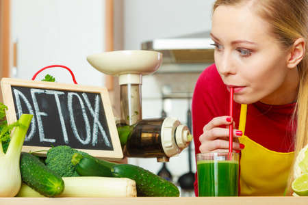 Drinks good for health, diet breakfast concept. Young woman in kitchen holding green healthy vegetable smoothie juice glass next to detox sign.の写真素材