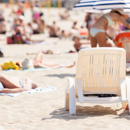 Sunbathing equipment concept. Deck chair on sandy beach with many people during summertimeの写真素材