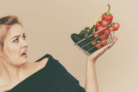 Adult woman do not like to eat vegetables, healthy food, vegetarian products. Female holding small shopping basket with green red vegetables, negative displeased face expression, on greyの写真素材