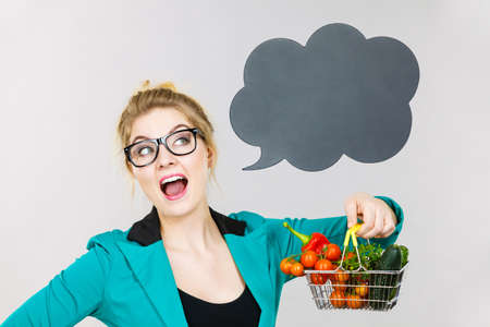 Buying healthy food, vegetarian products. Positive woman holding shopping cart with vegetables inside, standing near blank speech bubble with copy space for text, on greyの写真素材