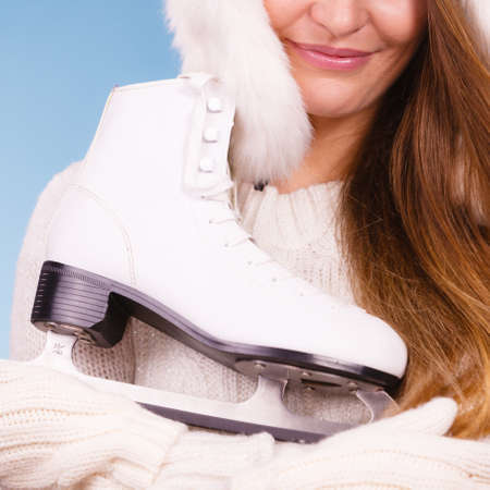 Winter sport activity concept. Girl wearing warm white clothes holding and hugging ice skate, blue background studio shot.の写真素材