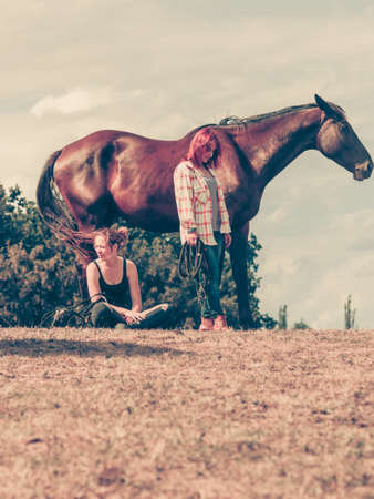 Animal and human love, equine concept. Jockey woman sitting relaxing with horses on meadow and western girl.の写真素材