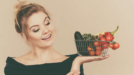 Buying good food, vegetarian products. Positive woman holding shopping basket with green red vegetables inside, recommending healthy high fibre diet, lifestyle modification, on greyの写真素材