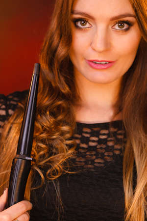 Haircare, hairstyiling at home concept. Portrait of young happy woman curling her long brown straight hair with curler. Studio shot on dark colorful backgroundの写真素材