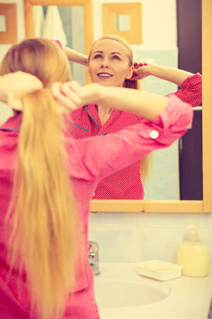 Woman combing brushing her long blonde smooth hair in bathroom, looking in mirror. Teen girl taking care refreshing her hairstyle in morning. Haircare concept.の写真素材