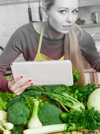 Young woman in kitchen having many green vegetables on table, holding tablet thinking about cooking something and searching for recipes in internetの写真素材
